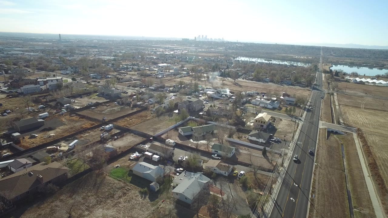 High aerial frame in Westminster, Colorado looking across houses, brown open parcels, a vertical road, water in the midground, and a hazy skyline, with no visible people.