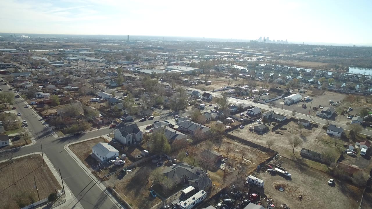 Drone-style view over Westminster, Colorado showing an intersection, residential yards, rows of houses, commercial rooflines, reflective water areas, and no visible people.