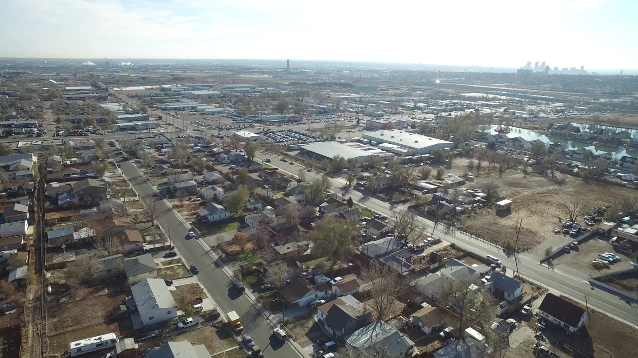 Aerial frame over Westminster, Colorado showing a straight residential street, small houses, warehouse roofs, reflective water in the distance, and no visible people.