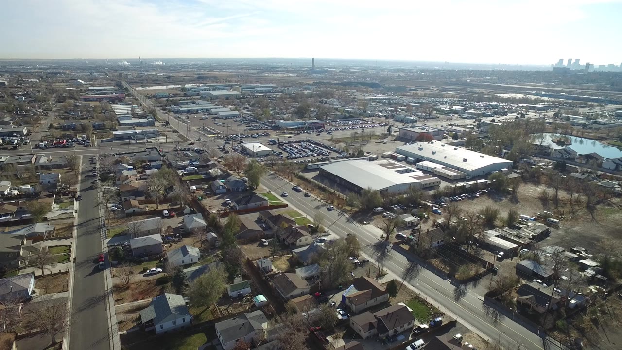 High drone-style frame in Westminster, Colorado showing a straight road corridor, small homes, large flat-roof industrial buildings, parked cars, and no visible people.