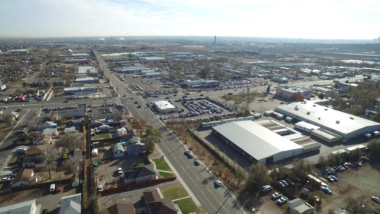 High aerial view over Westminster, Colorado with a long road, low industrial buildings, parking lots, scattered homes, leafless trees, and no visible people.