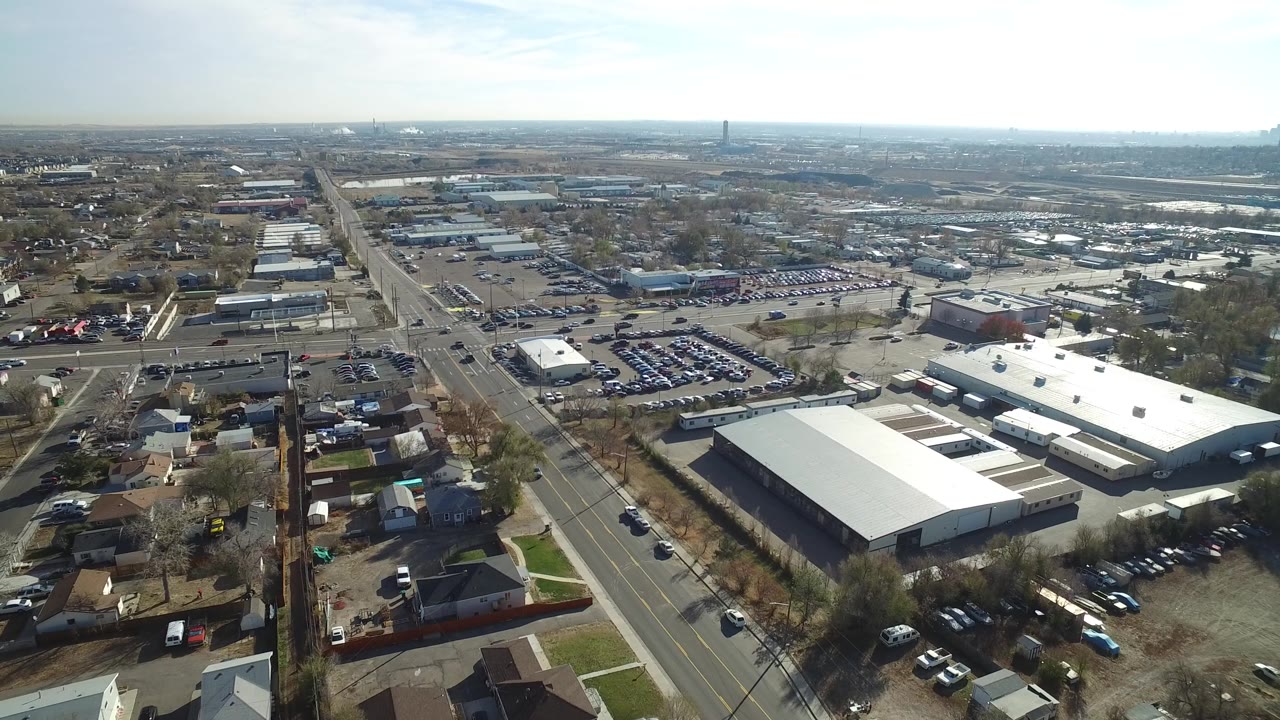 Aerial frame over Westminster, Colorado showing a main road leading toward the horizon, large white-roofed warehouses, parked cars, nearby homes, and no visible people.