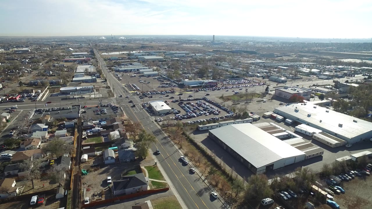 Drone view of a suburban commercial corridor with traffic, parking lots, homes, and warehouses.