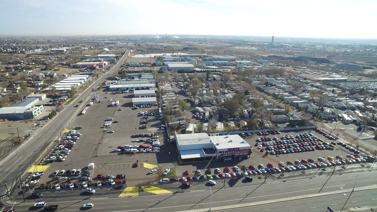 Opening aerial frame over Westminster, Colorado with a large parking lot in the foreground, commercial buildings and roads through the midground, and no visible people.