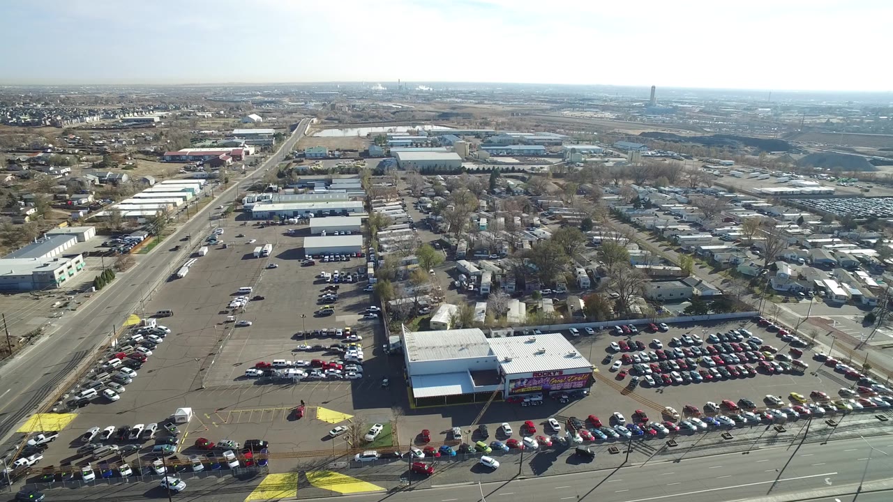 Drone-height view over a busy parking lot, broad street, small buildings, and open industrial land.