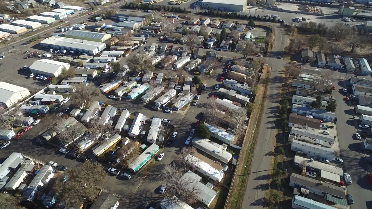 Aerial frame in Westminster, Colorado showing dense residential rows, parked cars along lots, a straight road on the right, and pale industrial roofs beyond, with no visible people.
