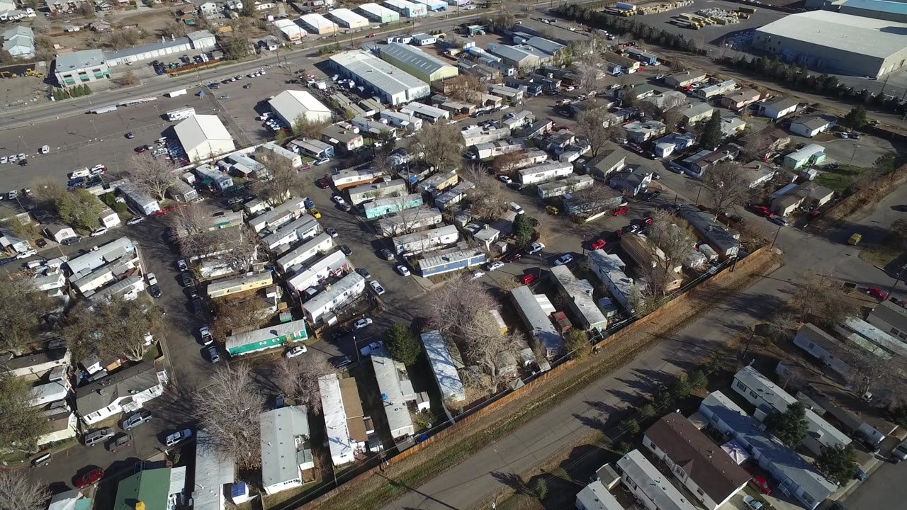 A high-angle aerial frame over Westminster, Colorado shows rows of mobile homes, parked vehicles, leafless trees, nearby roads, and industrial roofs in daylight, with no visible people.