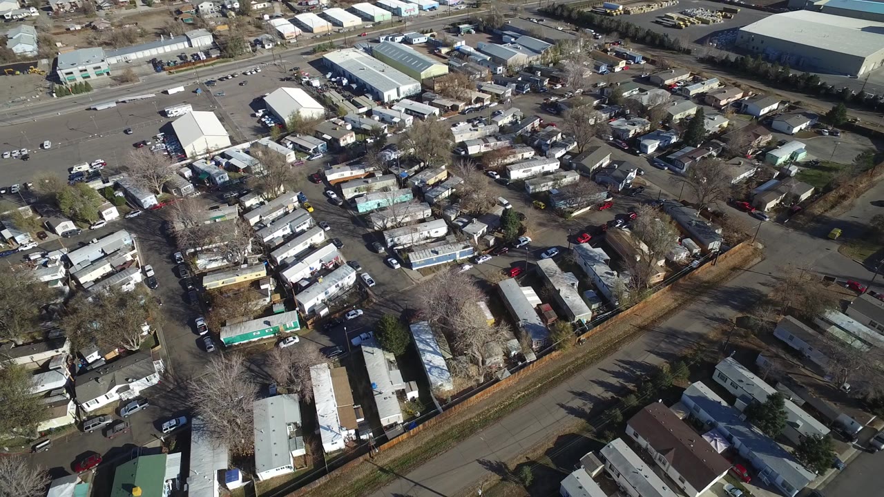 The second aerial frame keeps the Westminster neighborhood centered, with parallel mobile home roofs, service roads, parked cars, bare trees, and large warehouse structures beyond, with no visible people.