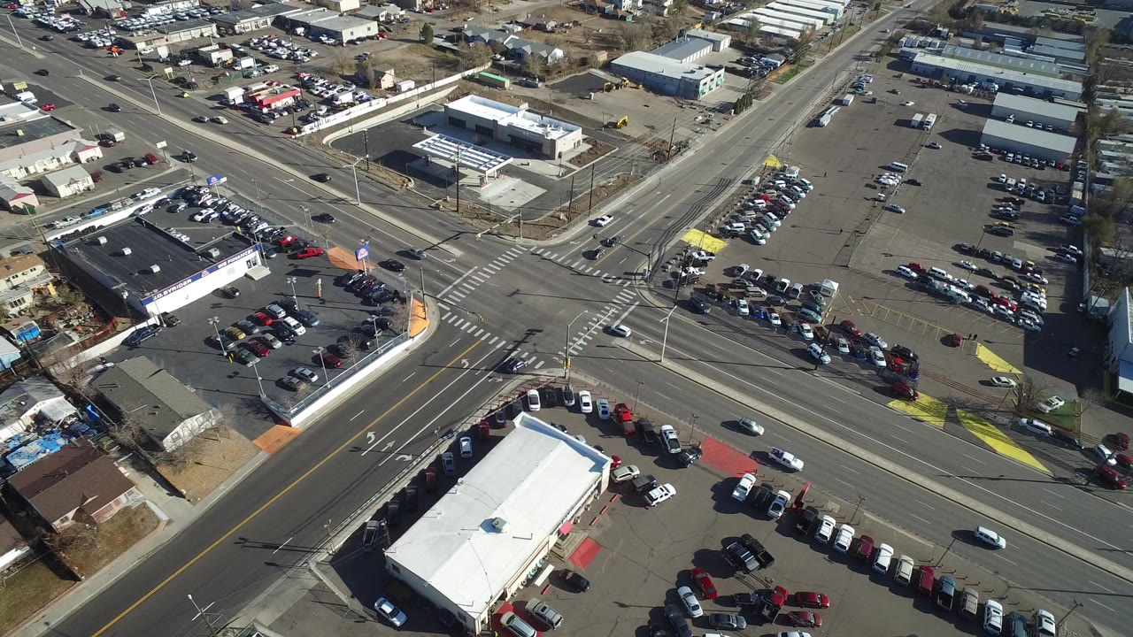 High aerial view over a Westminster, Colorado intersection with marked crosswalks, turning lanes, parked cars, low commercial buildings, and no visible people.