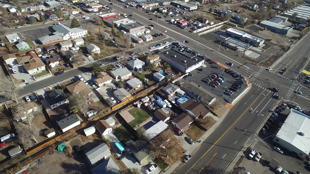 High-angle frame over Westminster, Colorado homes and backyards, with a diagonal road crossing the lower edge and a larger intersection beyond, with no visible people.