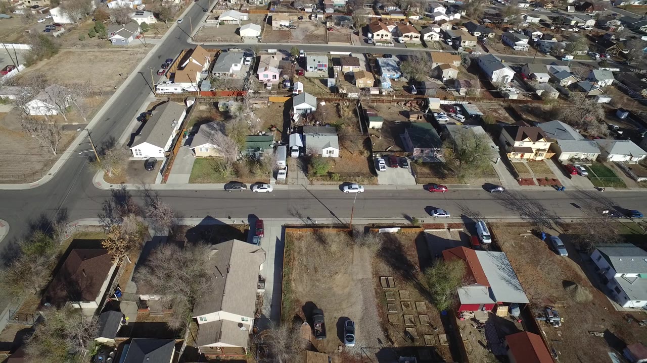Drone-height view of Westminster, Colorado residential blocks with a main road crossing the lower frame, parked vehicles, fenced yards, and no visible people.