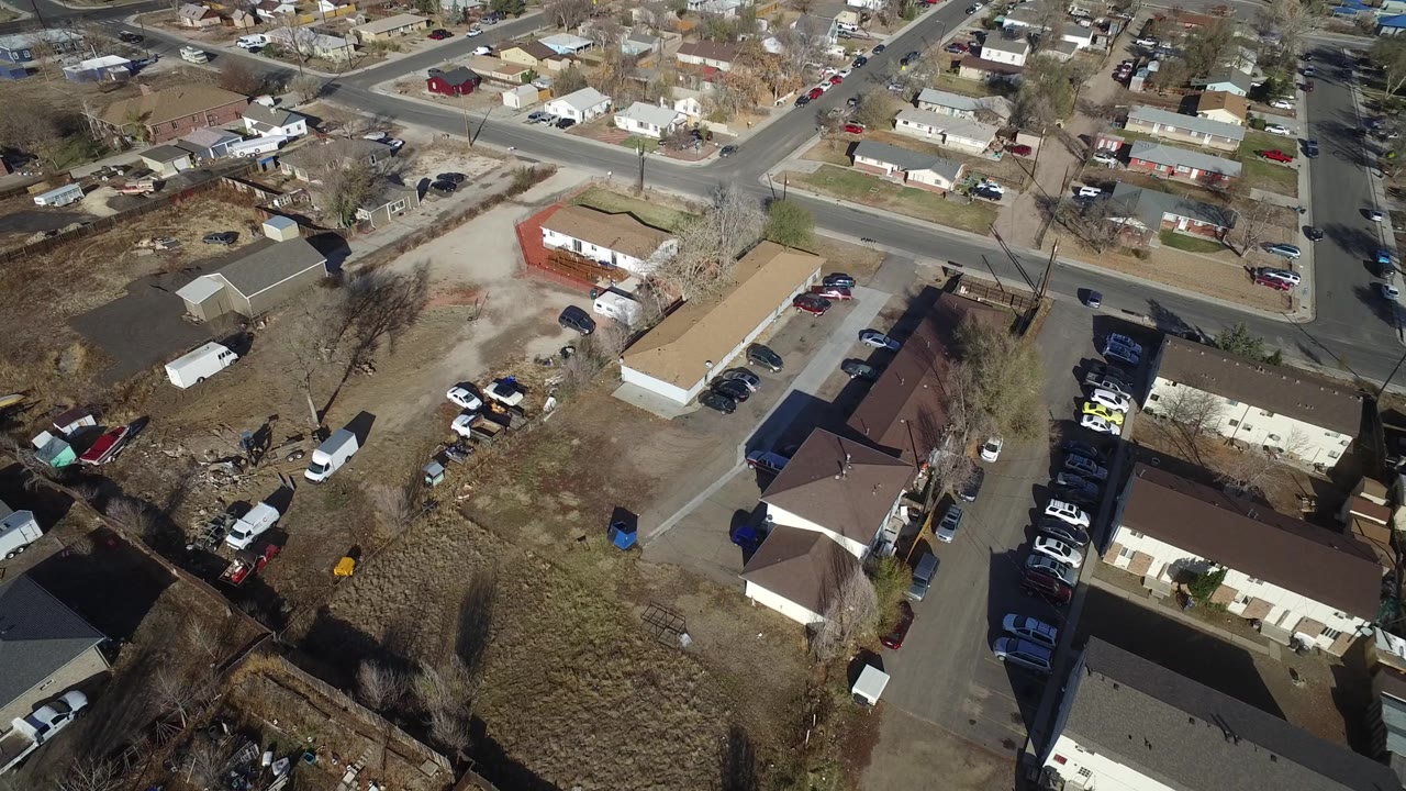 Aerial frame over Westminster, Colorado showing a road crossing the upper frame, residential roofs, open dirt patches, parked vehicles, and no visible people.