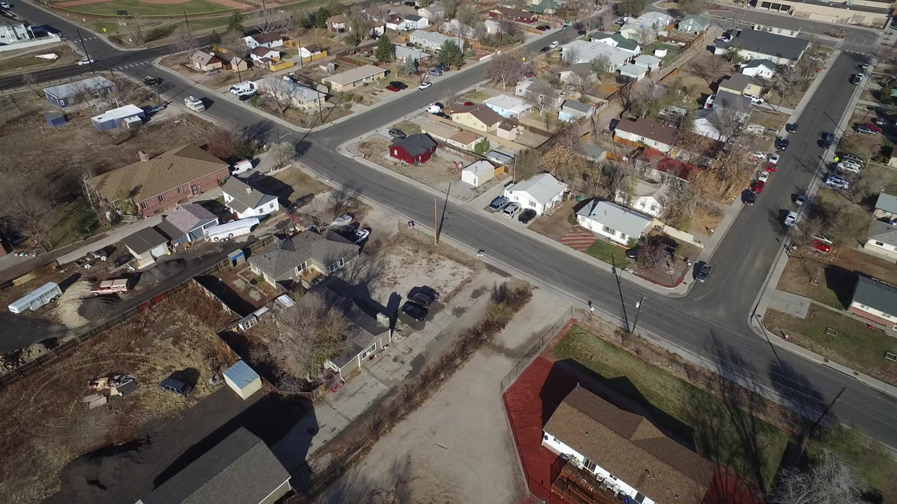 High aerial frame over Westminster, Colorado showing a broad residential intersection, detached houses, bare trees, parked vehicles, dirt lots, and no visible people.