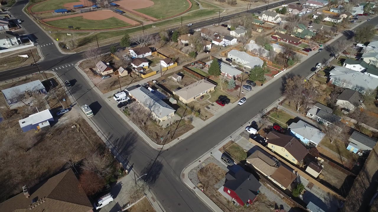 High overhead frame of Westminster, Colorado houses clustered around a quiet intersection, with baseball diamonds and park grass across the road, and no visible people discernible.