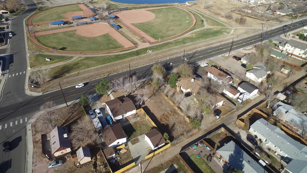 Aerial frame over Westminster, Colorado showing two green ball fields, a road with cars, nearby houses, bare trees, and no clearly visible people.