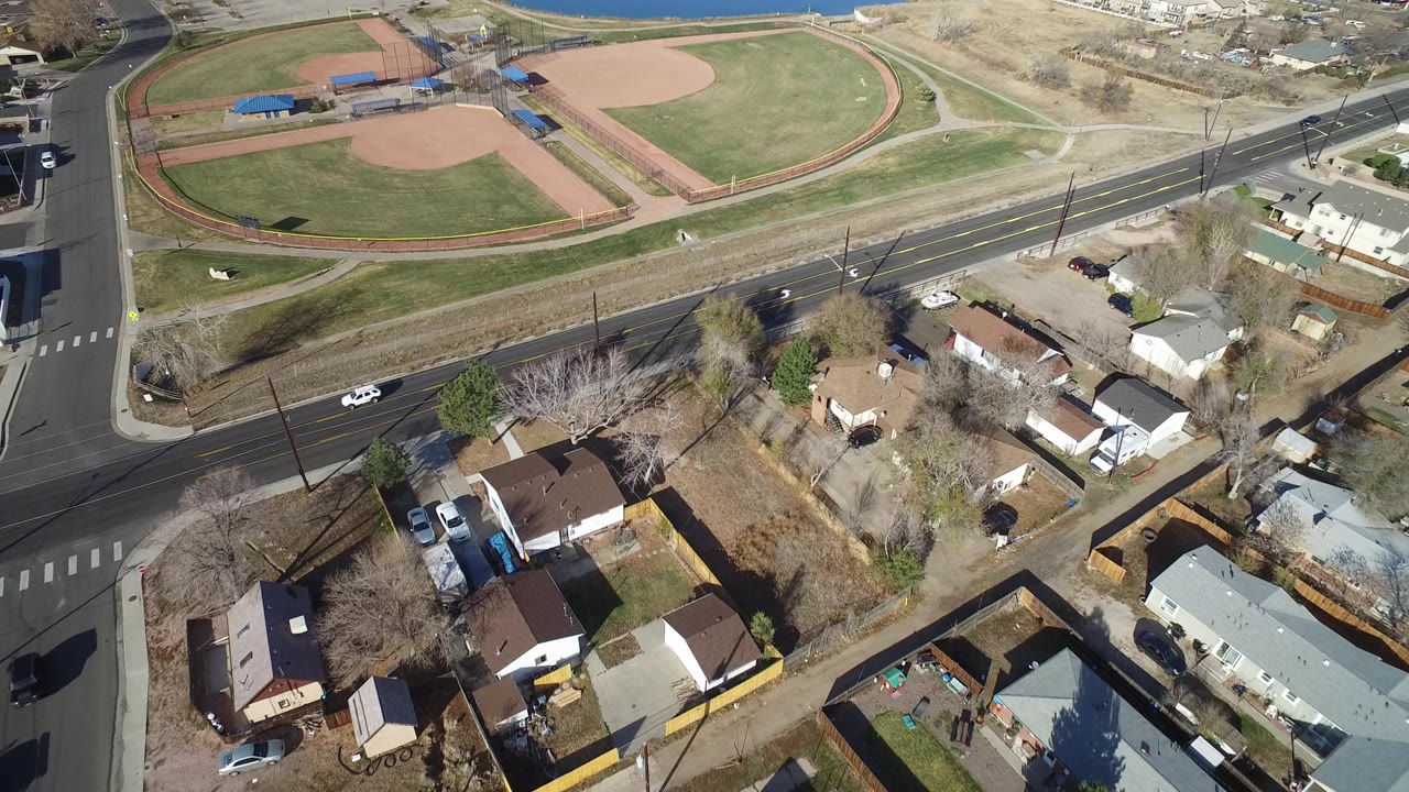 Aerial view in Westminster, Colorado with the baseball diamonds higher in frame, a blacktop road across the middle, parked and moving cars, private homes, and no clearly visible people.
