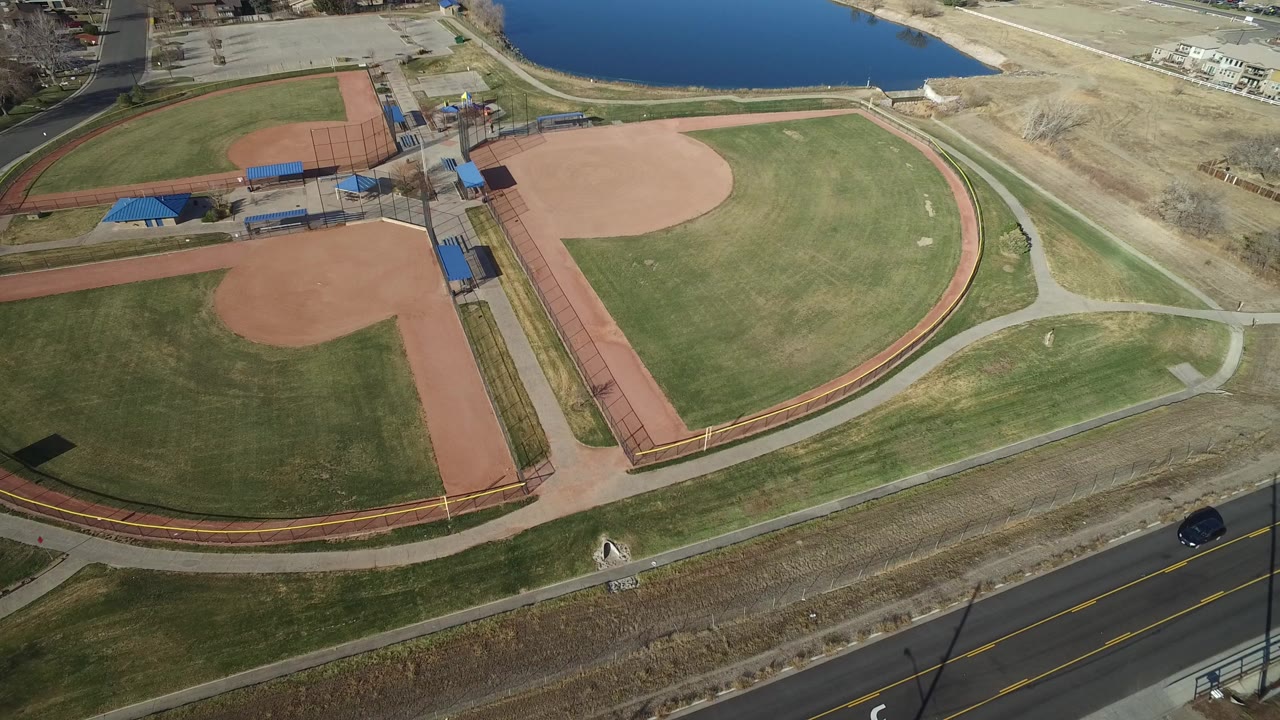 Overhead frame in Westminster, Colorado showing two ball fields, green grass, tan infields, blue-roofed structures, a road with a dark car, lake water, and no visible people.