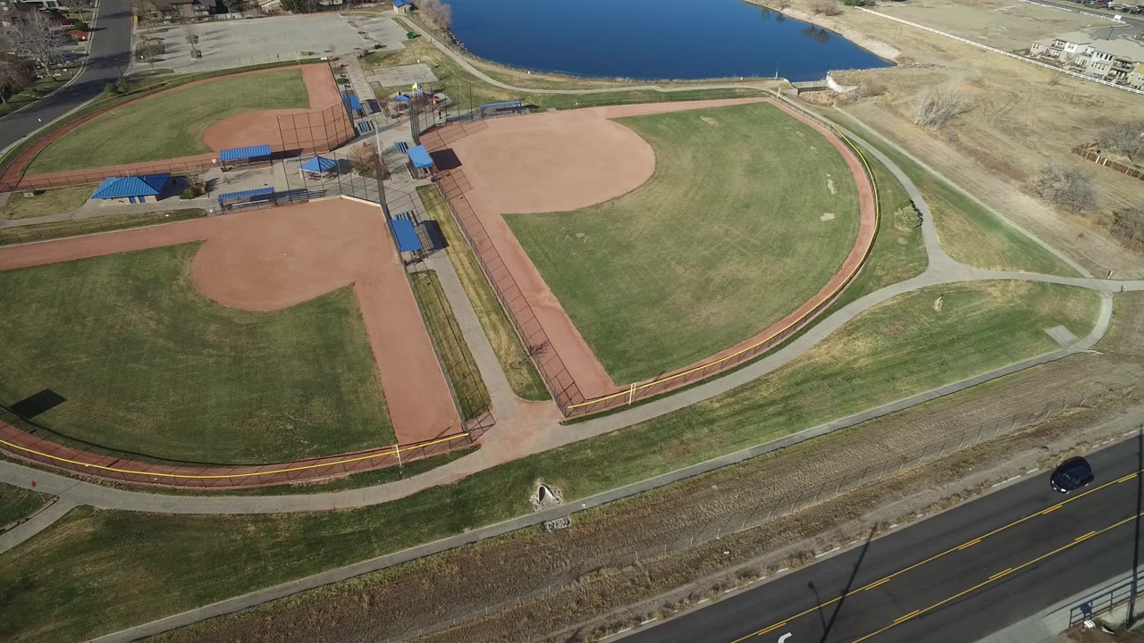 Aerial view in Westminster, Colorado with the main ball field centered, a second diamond at left, blue dugout roofs, curving paths, a two-lane road, lake shoreline, and no visible people.