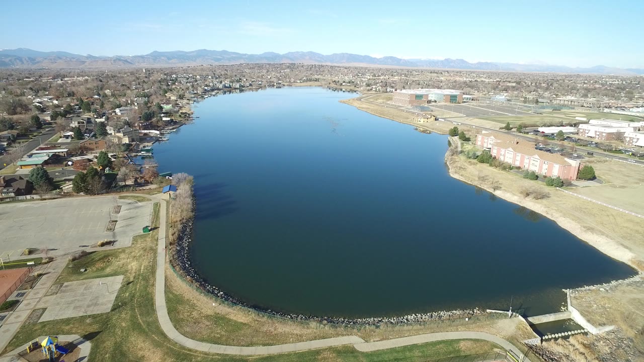 Aerial view of a blue lake bordered by paths, grass, homes, apartments, and distant mountains.