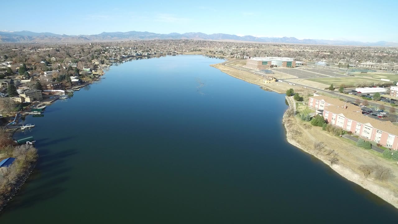 A high aerial frame in Westminster, Colorado centers the wide blue lake with the apartment-lined shoreline point at lower right, houses at left, recreation fields at upper right, and no recognizable people.
