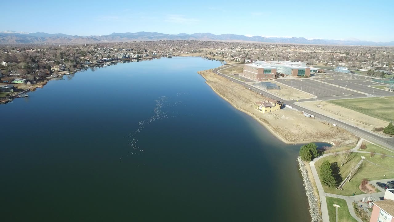 Aerial view over dark water toward a tan shoreline and suburban buildings.