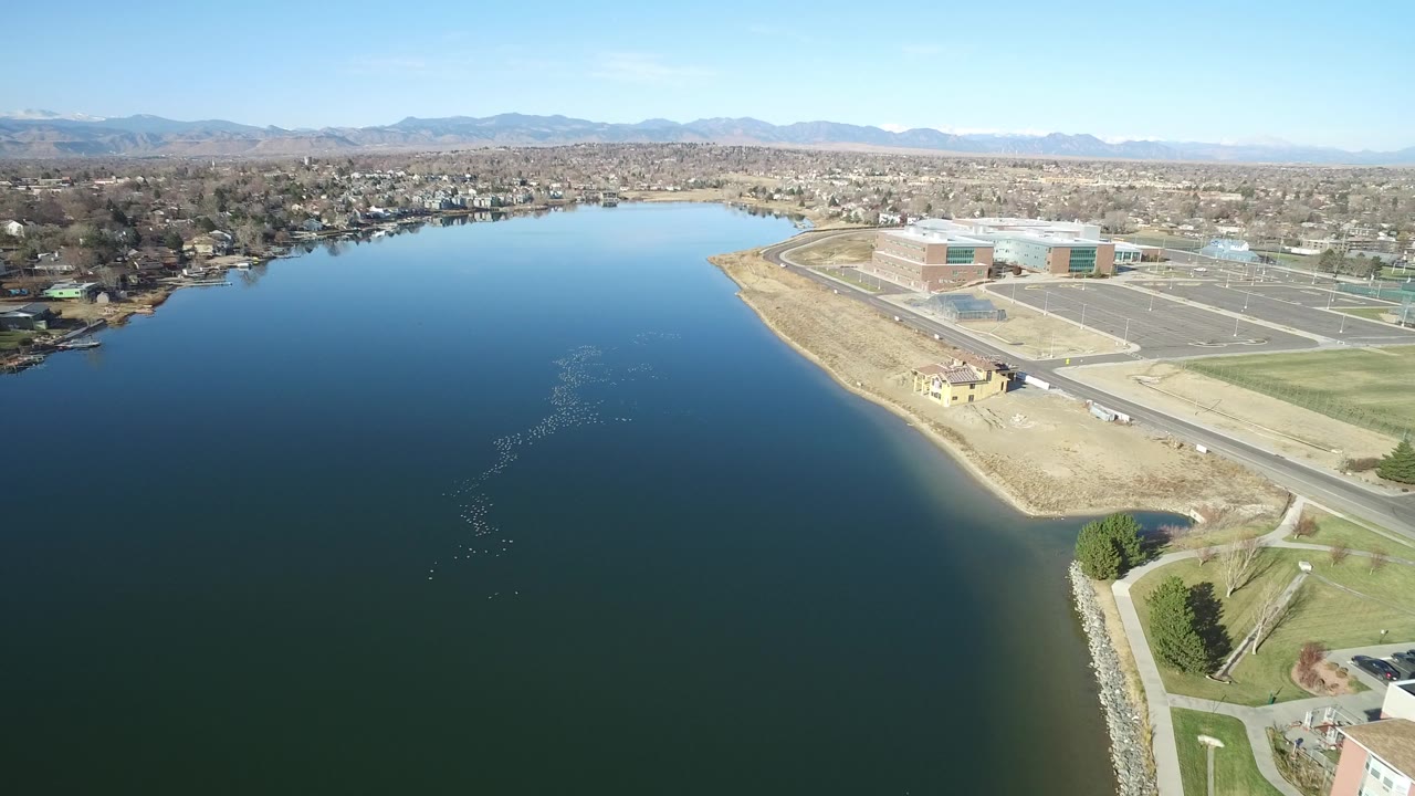 Aerial view in Westminster, Colorado with broad water in the foreground, a curved shoreline at right, empty paved lots, low institutional-looking buildings, mountains on the horizon, and no visible people.