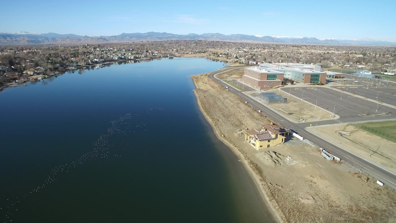 Aerial view over a lake beside a road, parking lots, low buildings, and distant mountains.
