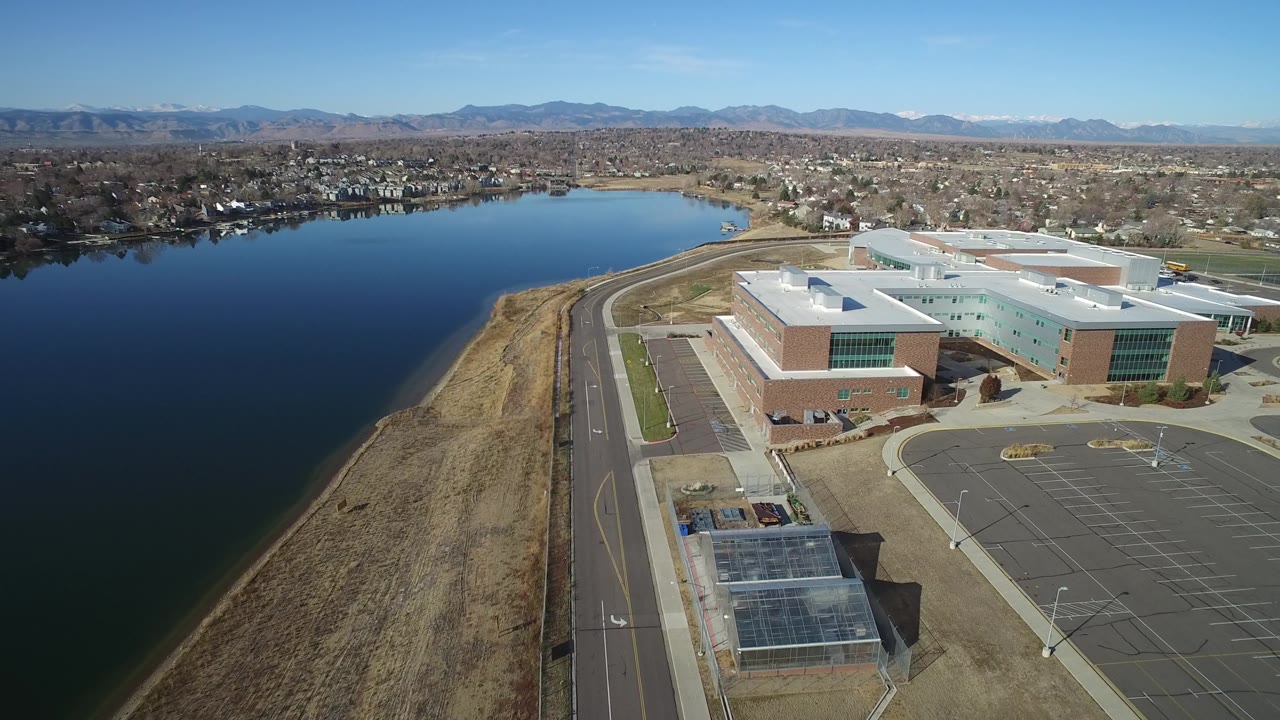 At 0:01, the aerial frame holds the Westminster office complex near the right side, with the lake filling the left half, a shoreline road below, residential blocks beyond the water, and no visible people.