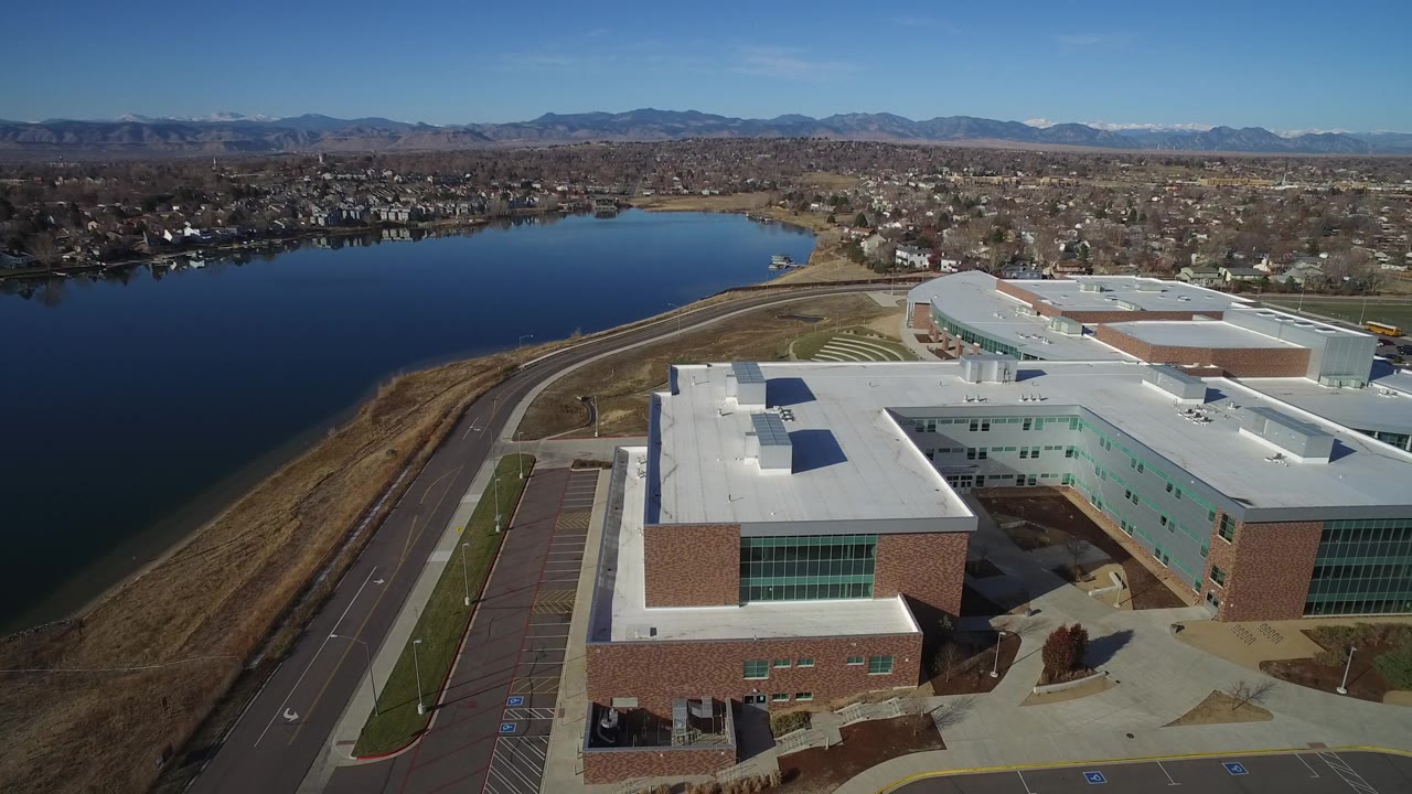 High aerial frame in Westminster, Colorado with the building roof centered low in frame, the lake to the left, residential blocks beyond, and no visible people.