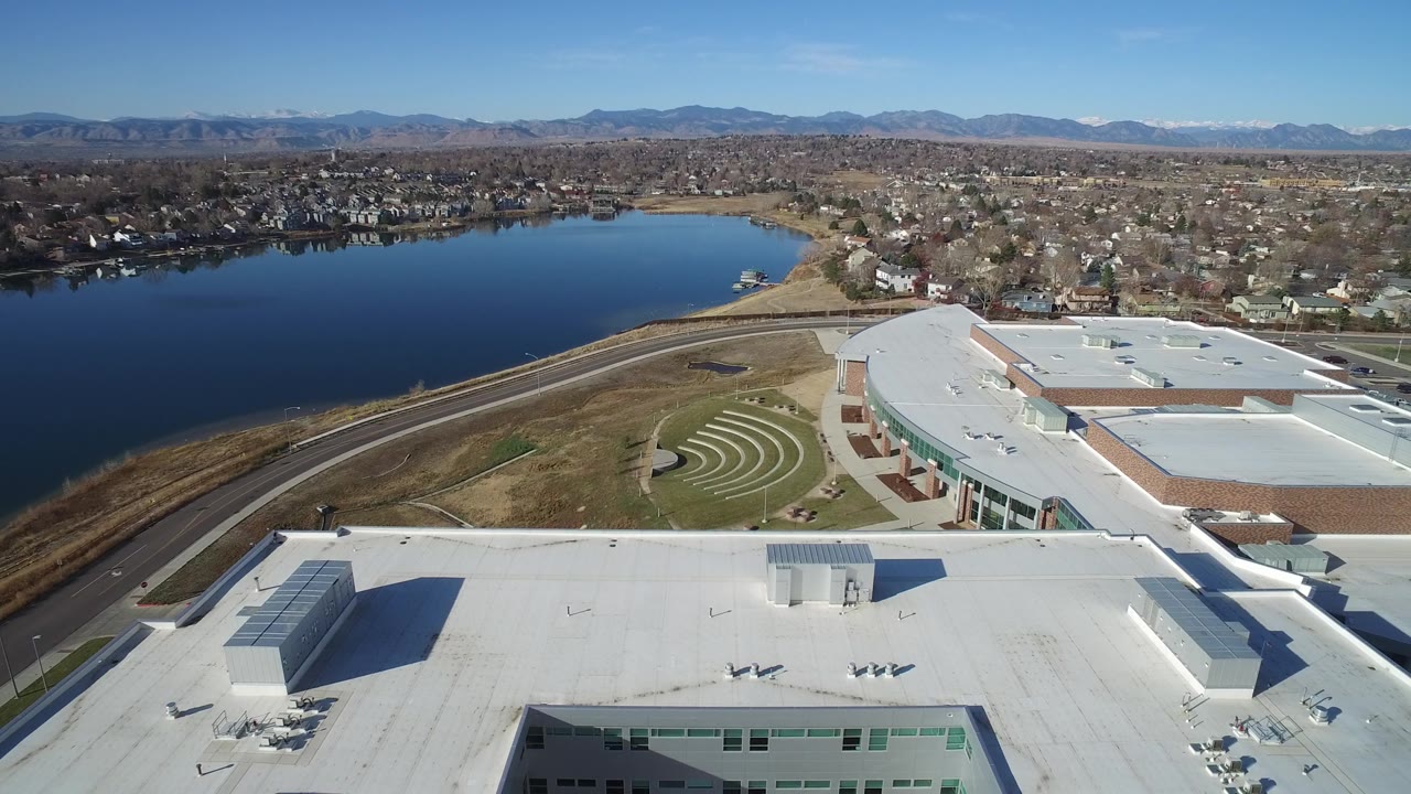 High aerial frame in Westminster, Colorado, showing the white roof closer across the lower foreground, the water curving along the left side, a road at the shore, dense homes in the distance, and no visible people.