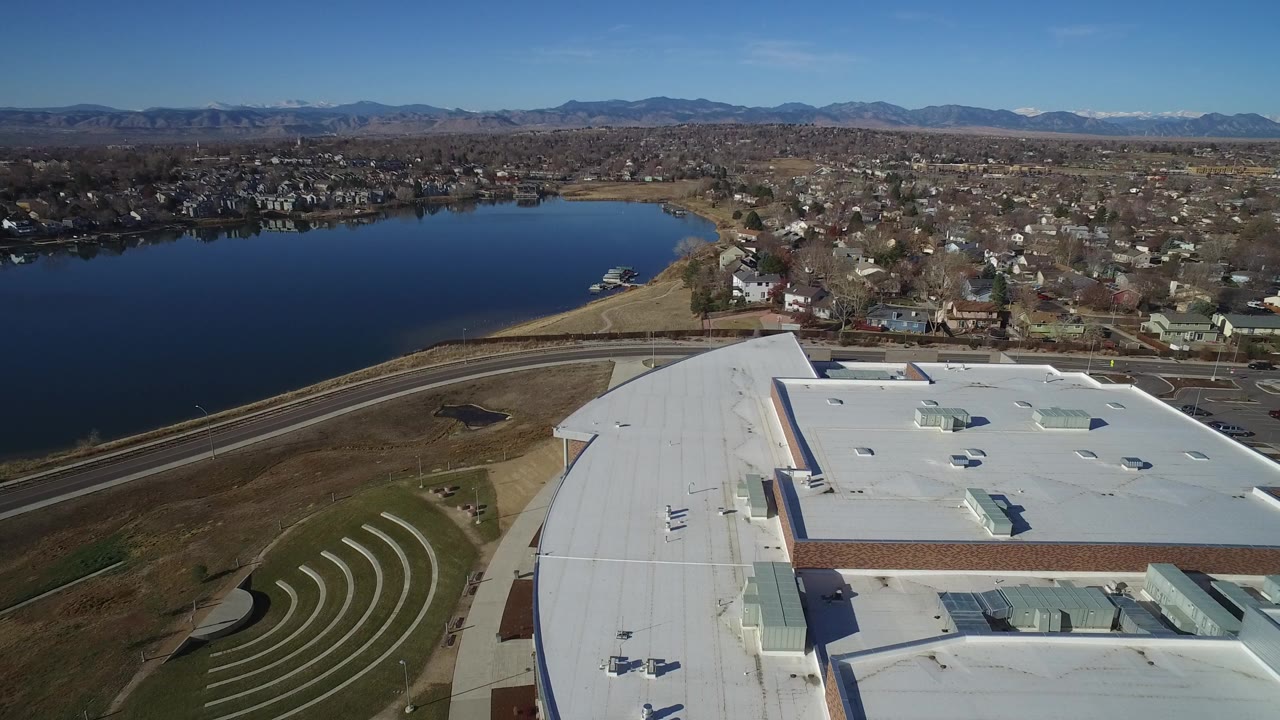 Opening aerial frame over Westminster, Colorado, showing a large white-roofed building in the foreground, a dark lake to the left, residential blocks beyond, distant mountains, and no visible people.