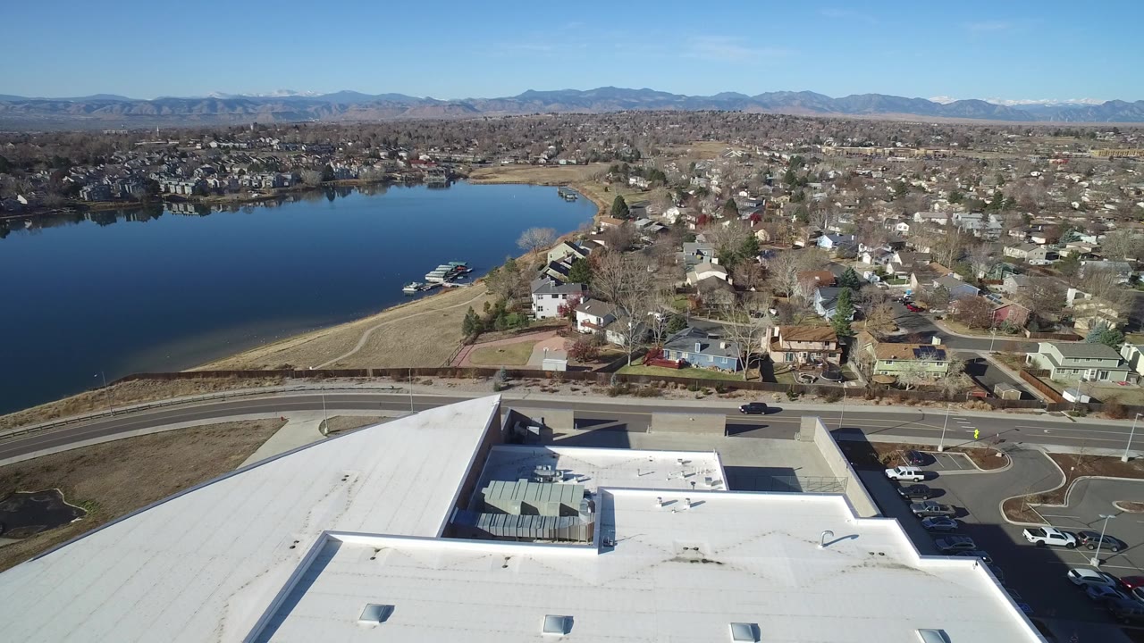 Aerial view over a flat white roof toward a lake, homes, roads, and mountains.