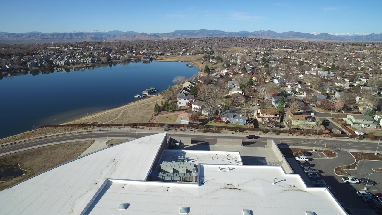 Westminster, Colorado aerial view with the lake filling the left side, a commercial-looking white roof below, a road with small vehicles, houses beyond it, and no visible people.