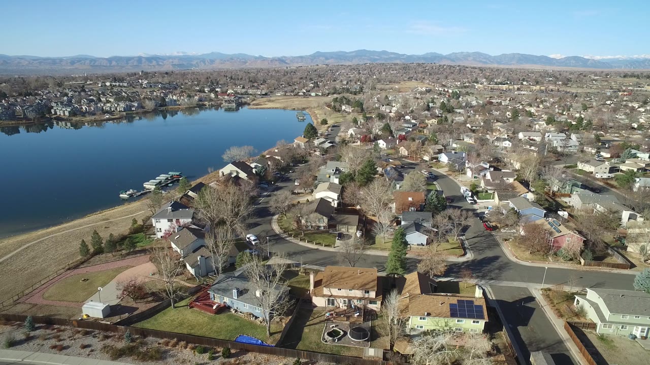 Westminster, Colorado aerial view one second in, with the lake filling the left side, tightly spaced homes and yards on the right, mountains along the horizon, and no visible people.