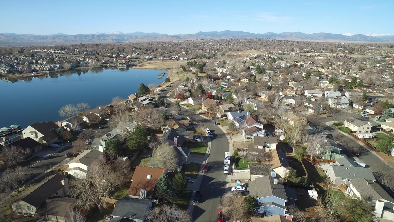 Aerial frame looking across Westminster, Colorado residential blocks, with the lake on the left edge, parked vehicles along streets and driveways, and no visible people.