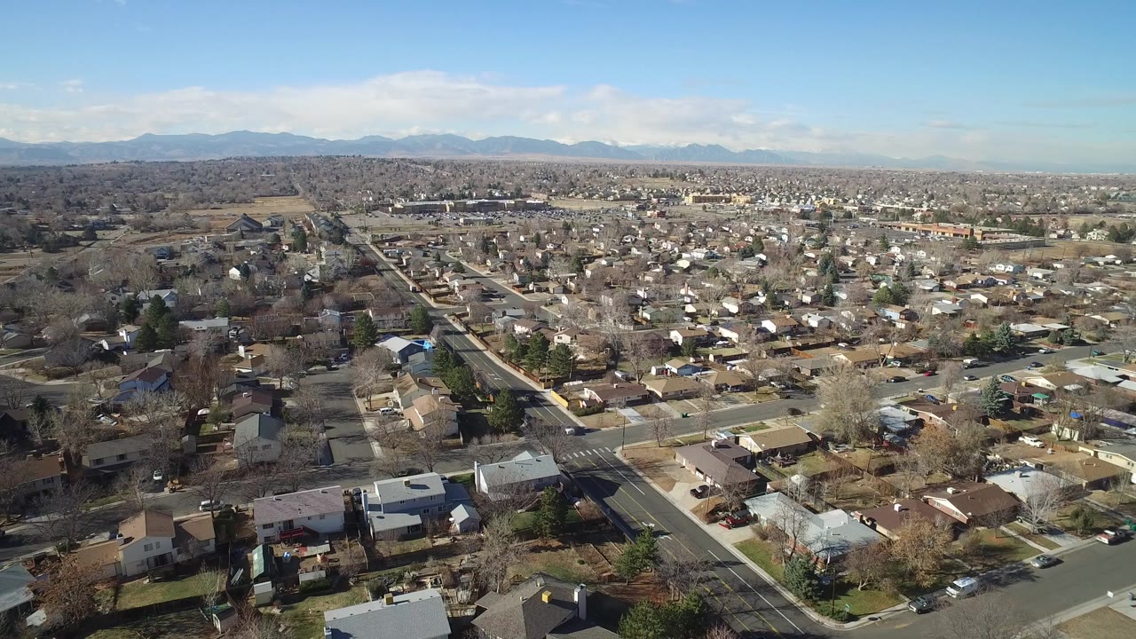 Aerial view over a residential neighborhood with roads, rooftops, open land, and distant mountains.