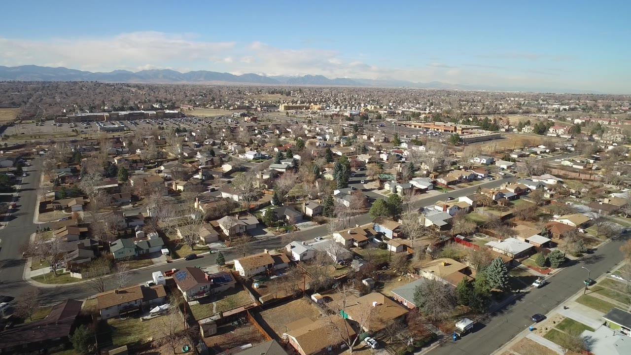 A wide drone frame over Hidden Lake, Colorado shows residential blocks, leafless trees, small yards, parked cars, and mountains along the background, with no visible people.