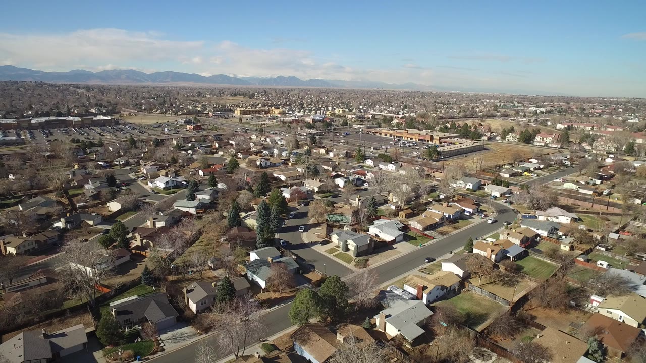 Elevated view over Hidden Lake, Colorado homes, yards, and curving residential streets under a blue sky, with distant mountains and no clearly visible people.
