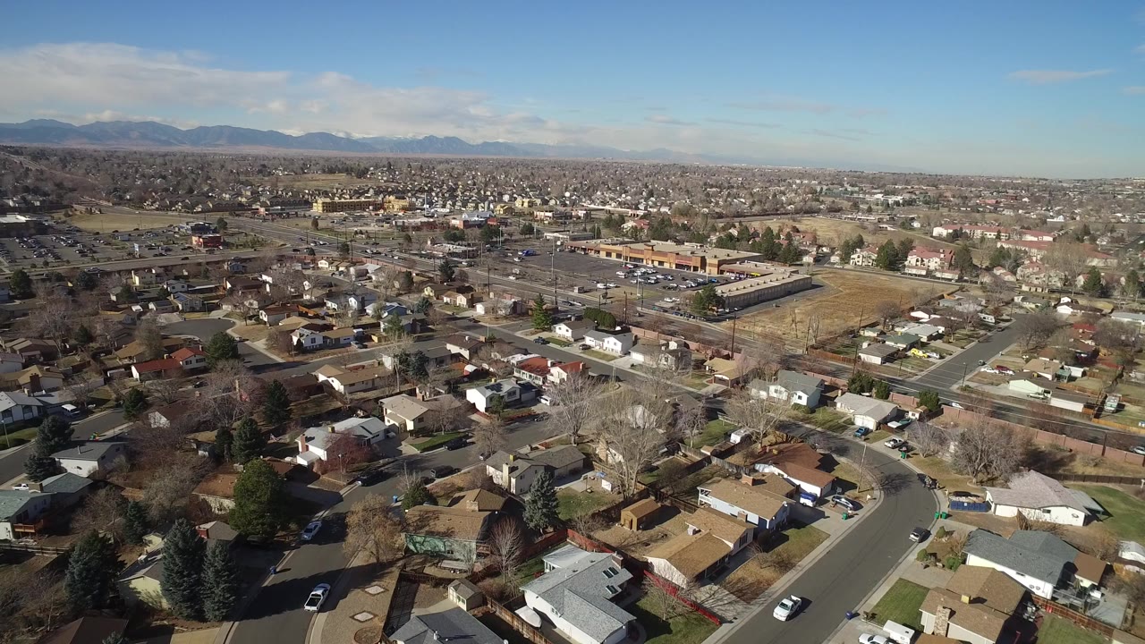 High aerial view over Hidden Lake, Colorado, showing residential rooftops, curving streets, bare trees, open lots, distant mountains, and no visible people at the start of the clip.
