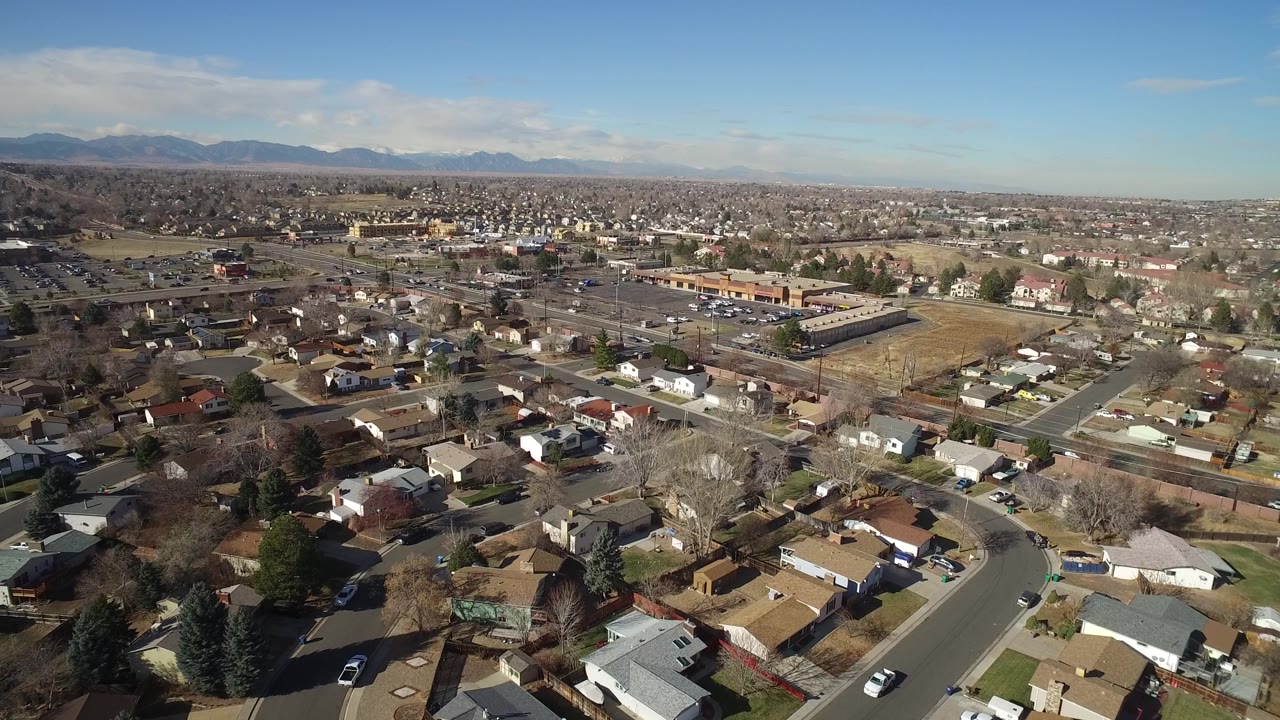 Aerial frame over Hidden Lake, Colorado, with houses in the foreground, a larger tan building near the middle distance, road traffic, open ground, distant mountains, and no visible people.