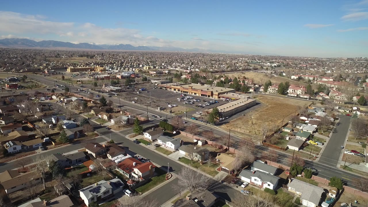 Aerial frame over Hidden Lake, Colorado showing foreground houses, a broad road with small cars, a parking lot, low businesses, open grassland, and no recognizable faces.
