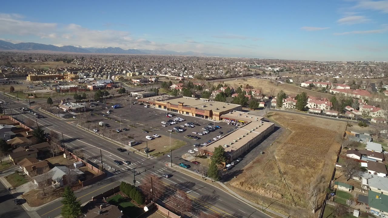 Opening aerial frame over Hidden Lake, Colorado with a commercial strip building and parking lot in front, homes and open ground beyond, distant mountains on the horizon, and no visible people.