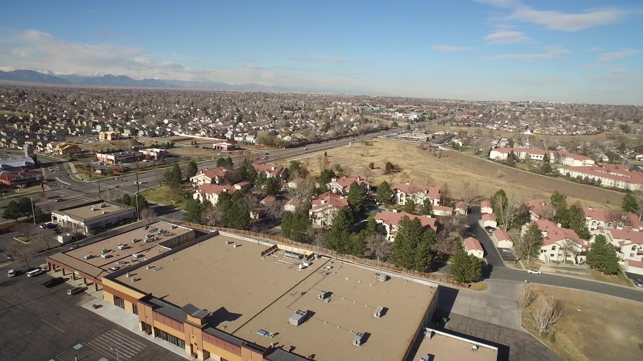 Aerial view over commercial rooftops, red-roofed homes, roads, open fields, and distant mountains.