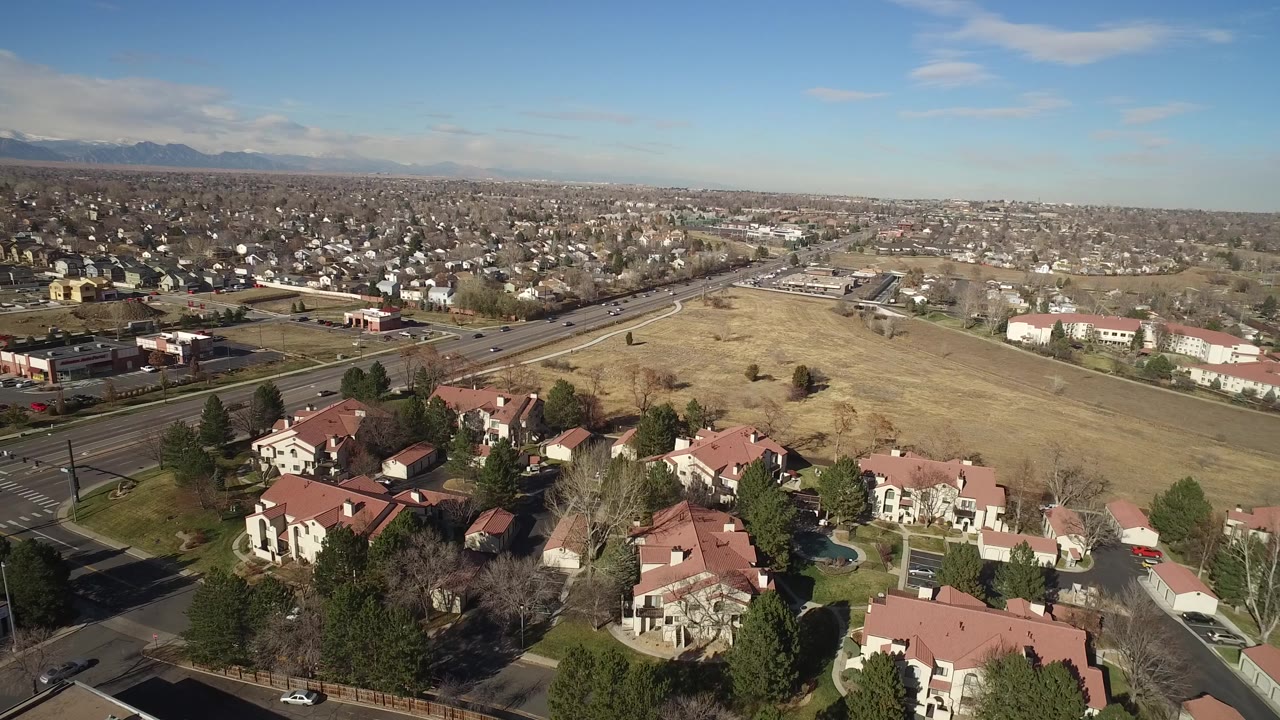 Aerial frame over Hidden Lake, Colorado showing clustered homes at the lower edge, a road crossing the scene, a dry field in the center, distant neighborhoods, mountains, and no visible people.