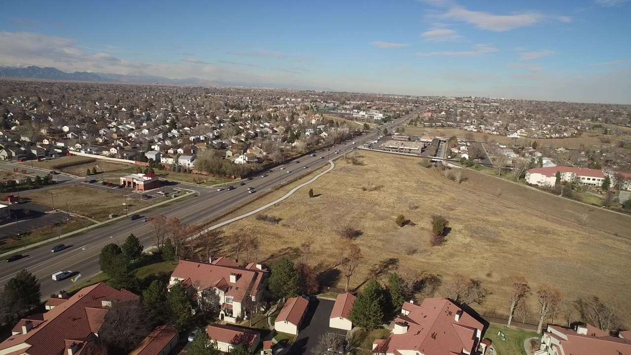 Opening aerial view in Hidden Lake, Colorado, looking over red-roofed homes, a dry open field, a multi-lane road with light traffic, distant neighborhoods, mountains, and no visible people.