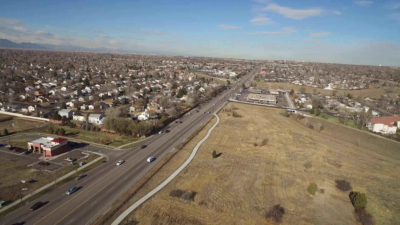 Aerial frame in Hidden Lake, Colorado showing cars on a straight arterial road, an open brown field to the right, houses to the left, and no visible people.