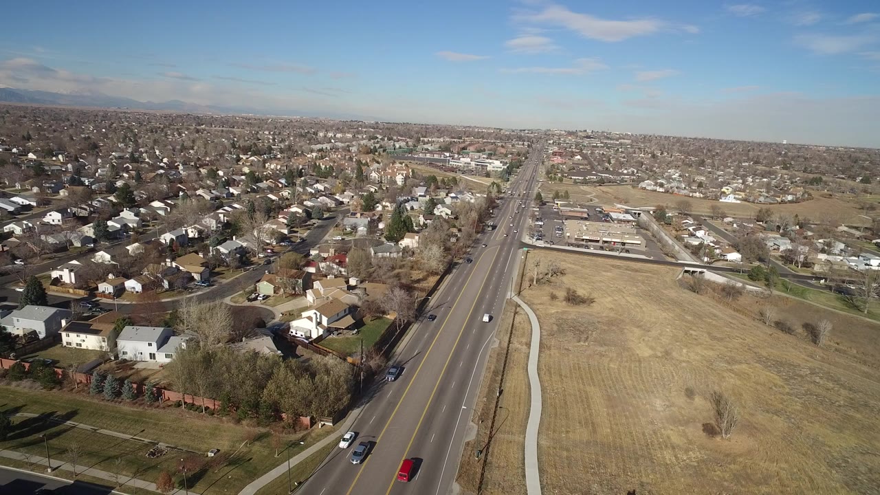 High aerial frame over Hidden Lake, Colorado showing a four-lane road running toward the horizon, houses on the left, tan open field on the right, moving cars, and no visible people.