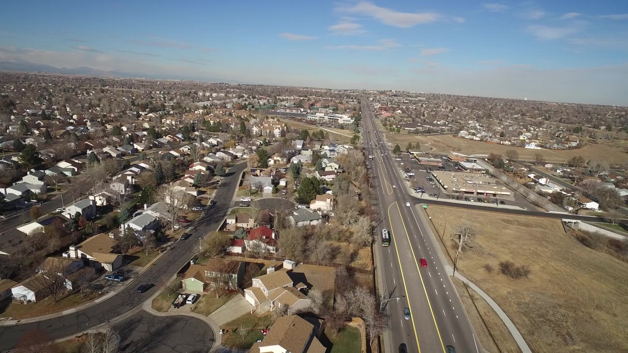At 0:01, the Hidden Lake, Colorado aerial keeps the divided highway near the center right while houses, leafless trees, parked vehicles, and open lots fill the foreground with no visible people.