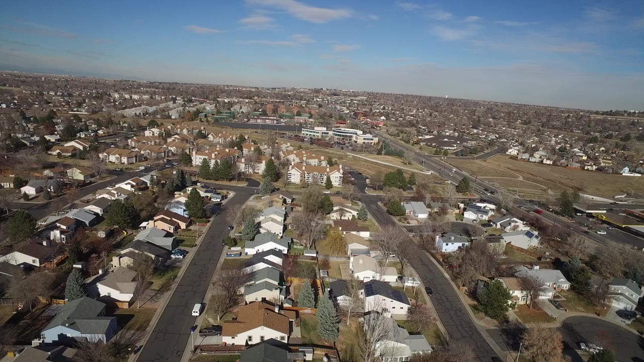 High aerial view of suburban houses, roads, open land, and distant neighborhoods under blue sky.