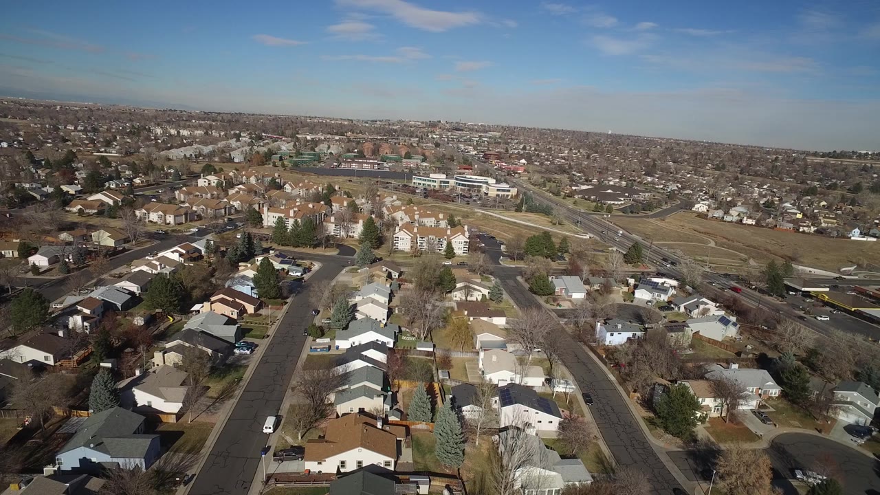 One second into the clip, the Hidden Lake, Colorado aerial view centers a straight neighborhood street with houses below, open land to the right, and no visible people.